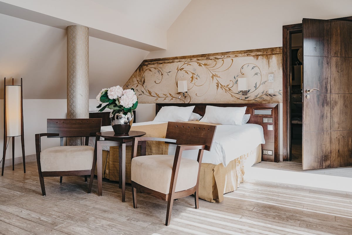 Une chambre d’hôtel comprenant un lit, deux chaises, une table, un vase avec des fleurs et une tête de lit décorative.