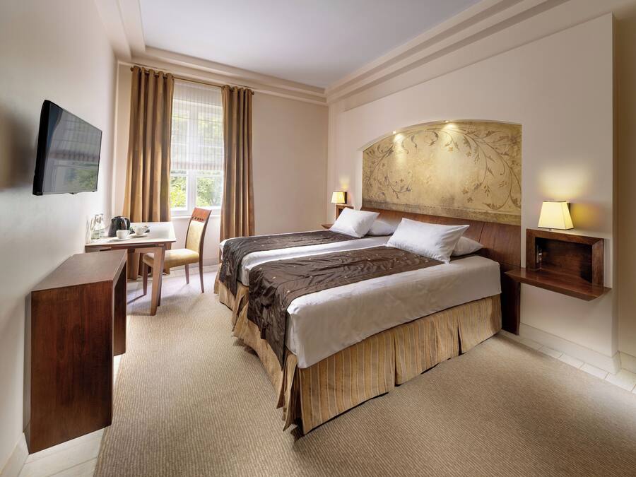Ein Hotelzimmer mit Bett, Nachttisch, Stuhl, Schreibtisch, Fernseher und einem fenster mit Vorhängen.