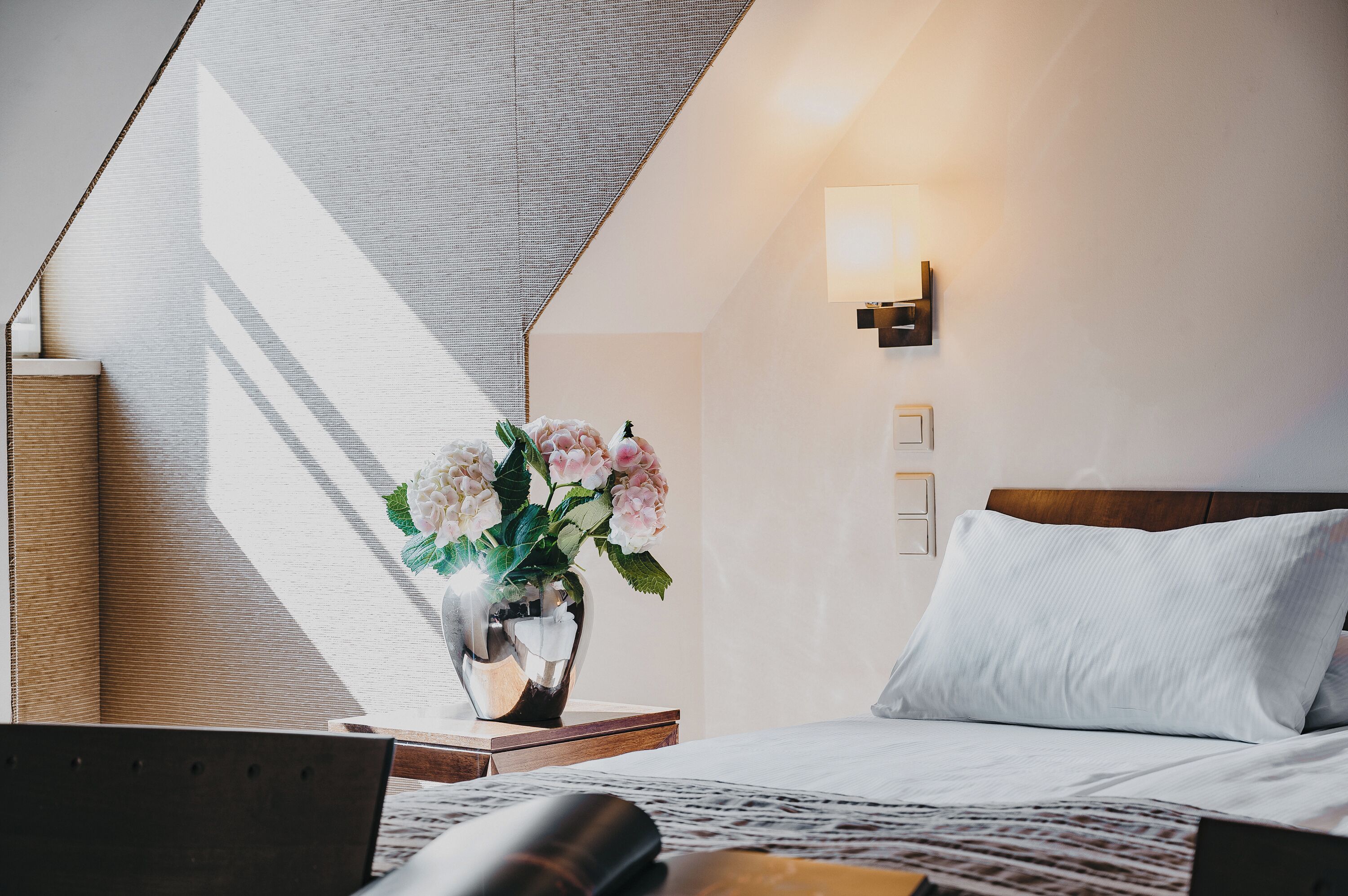 A hotel room with a bed, a bedside table with a vase of flowers, a lamp, and a view of the outside.