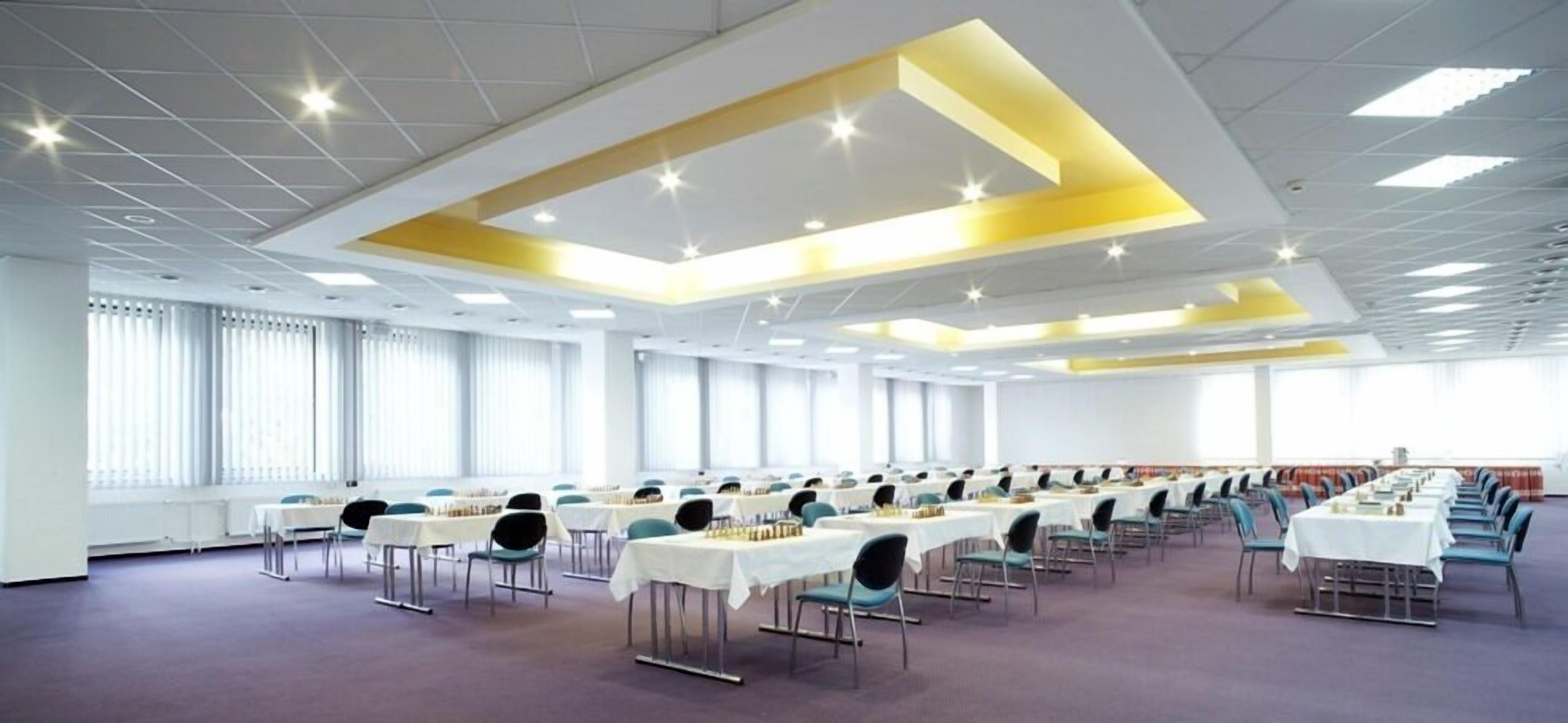 A conference room with rows of tables and chairs, large windows, and a modern ceiling design.