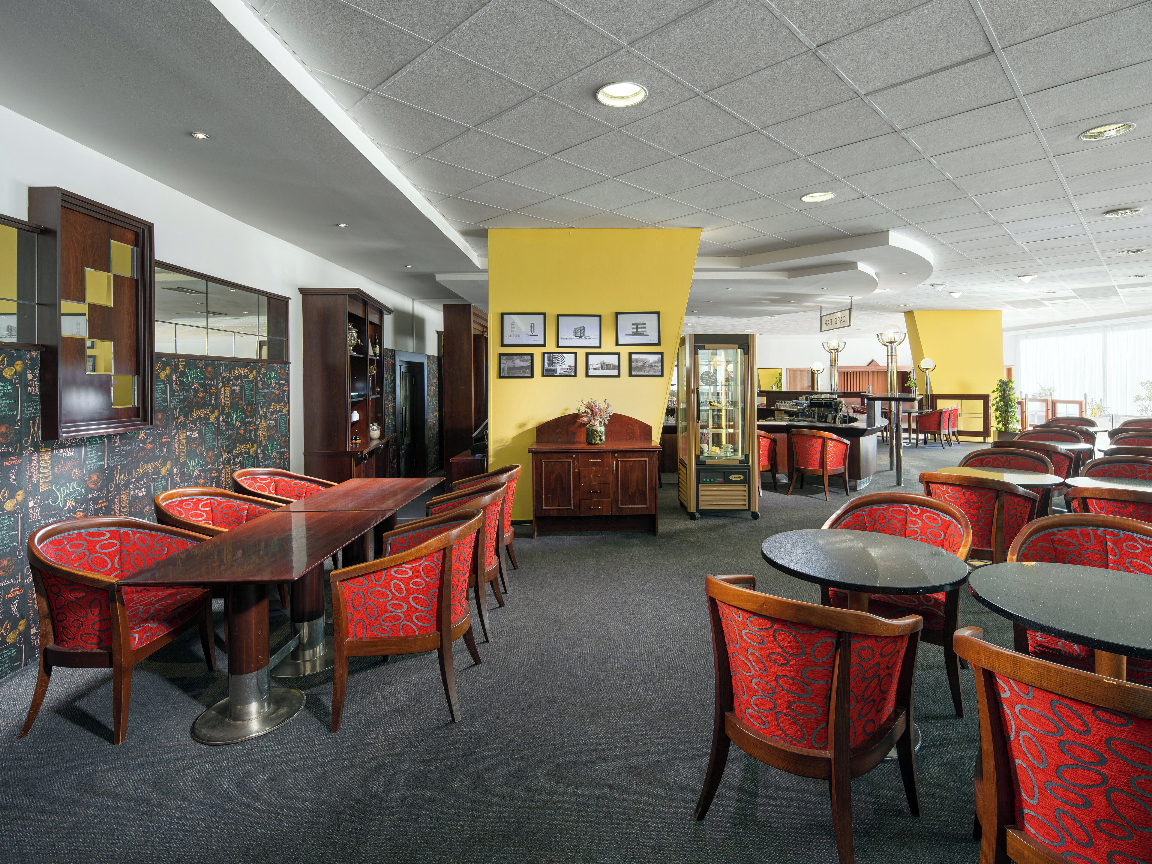 A restaurant with wooden tables and red patterned chairs, a yellow accent wall, and a bar area.