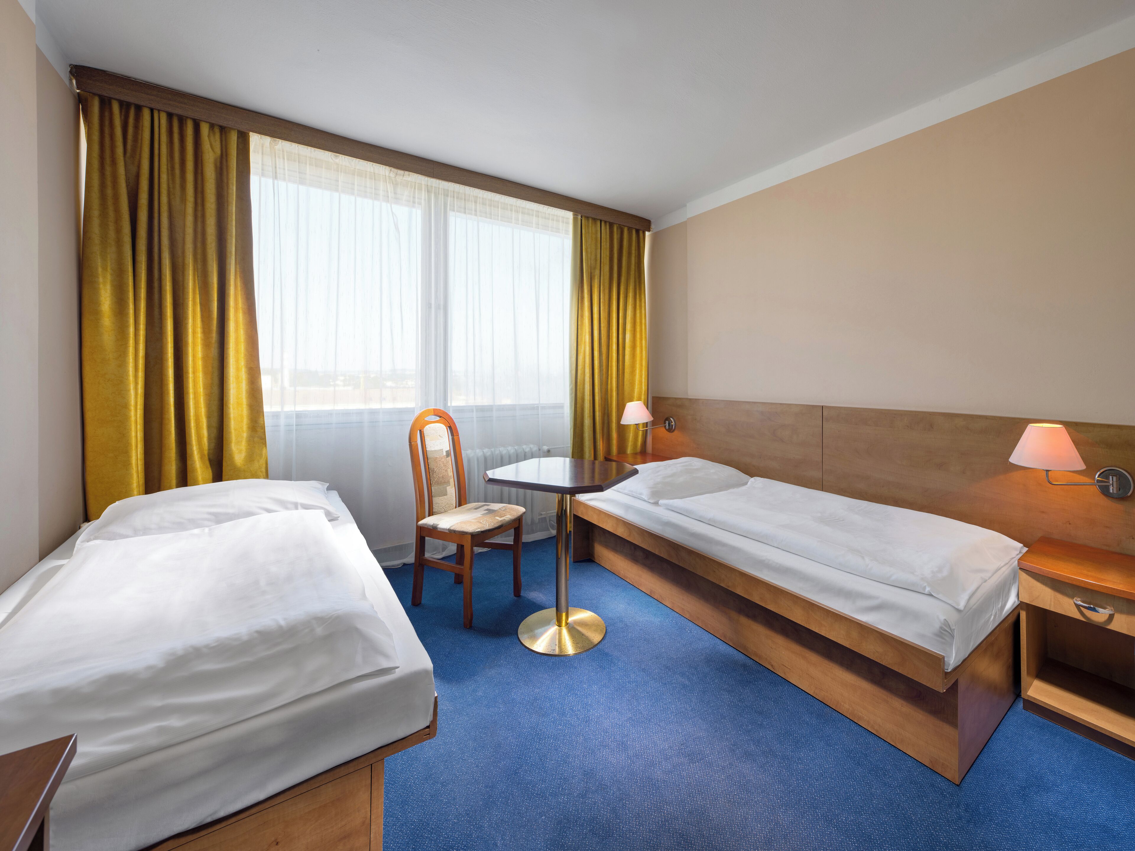 A hotel room with two single beds, a small table, a chair, and a lamp.