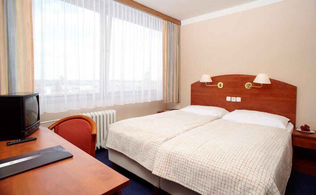 Een hotelkamer met een bed, een bureau met een computer, een stoel en een raam met gordijnen.