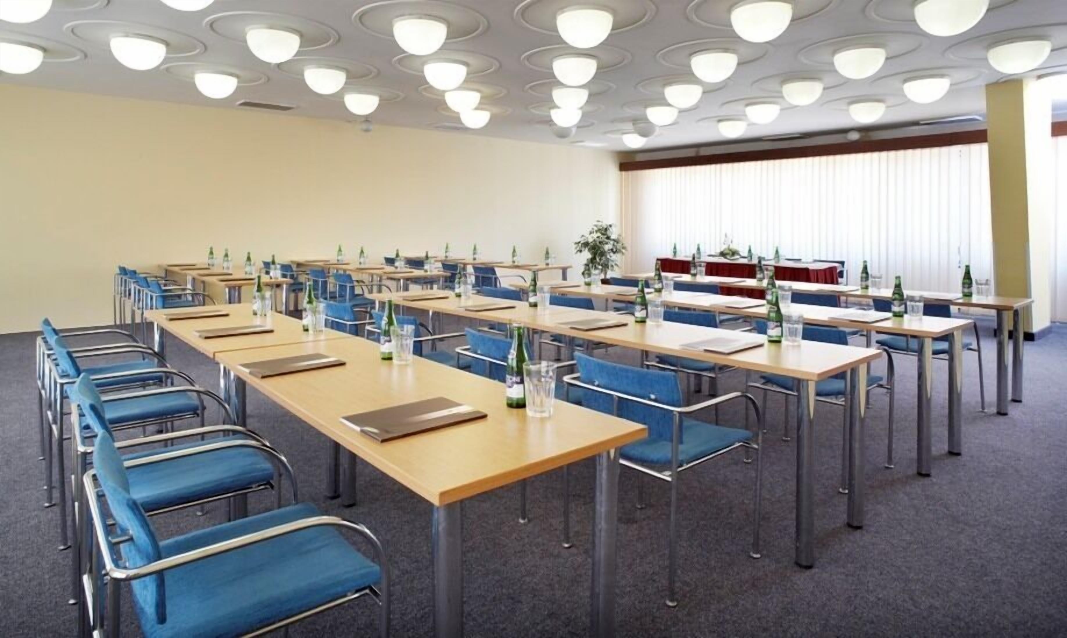 Une salle de conférence avec des rangées de tables et de chaises, équipée de bouteilles d’eau et de verres.