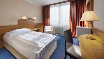 Une chambre d’hôtel comprenant un lit, un bureau, deux chaises, une lampe, une fenêtre avec des rideaux et une lampe fixée au mur.