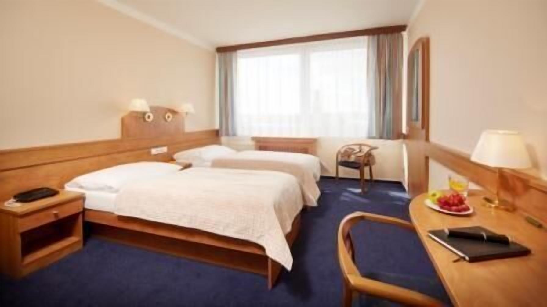 A hotel room with two beds, a desk with a chair, a small table with a plate of fruit, and a window with curtains.