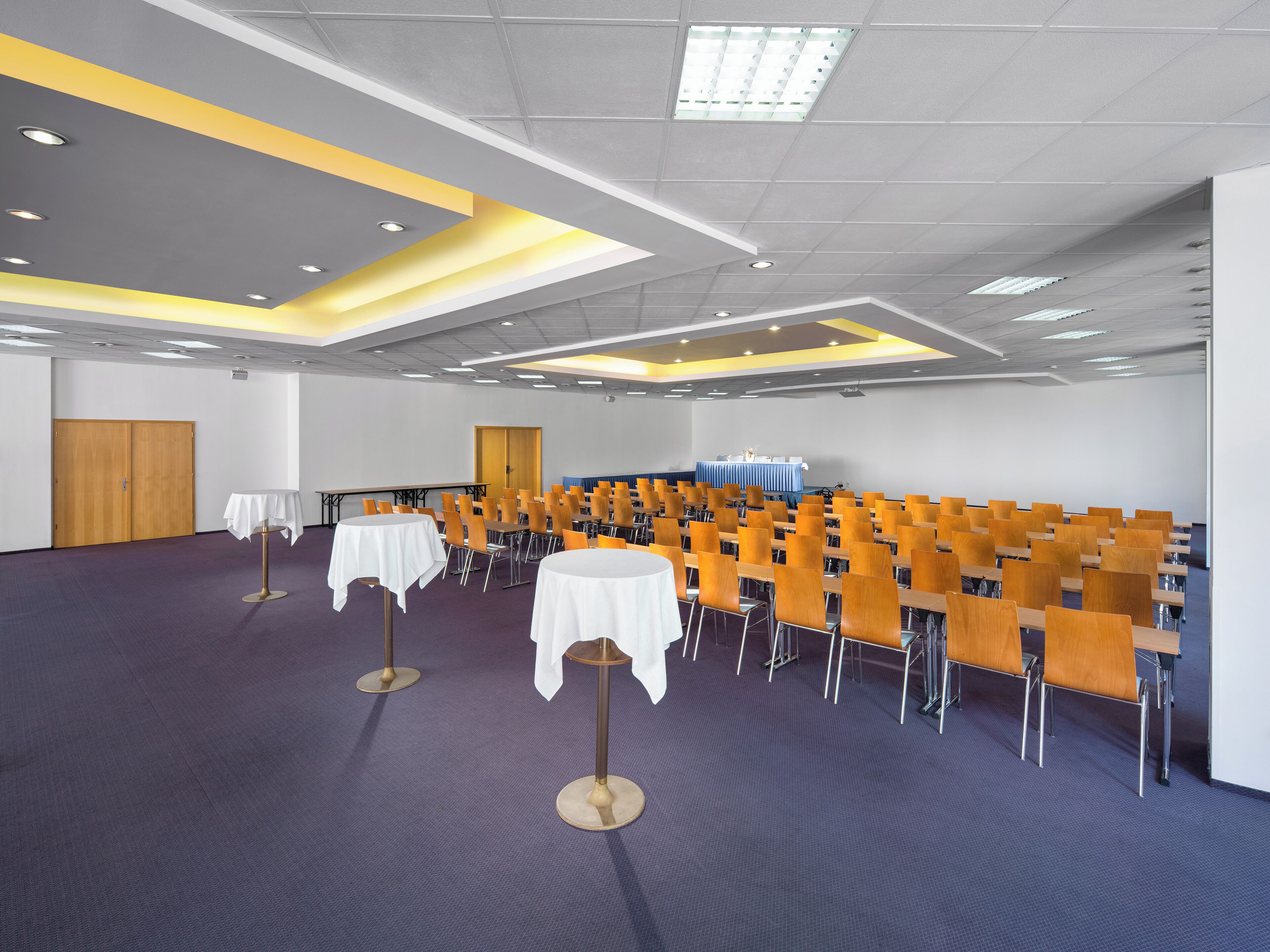 A conference room with rows of chairs and tables, a stage with a blue backdrop, and a ceiling with recessed lighting.