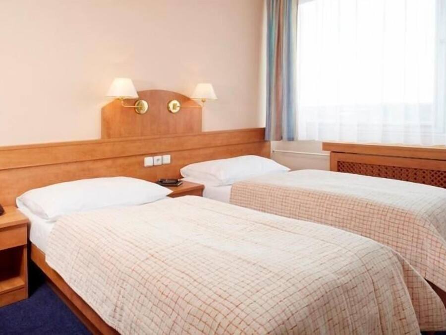 A hotel room with two single beds, wooden headboards, bedside lamps, a nightstand, and a window with curtains.