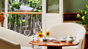 A room with a view of trees through a window, a chair, a table with a teapot, cups, and a vase with flowers.