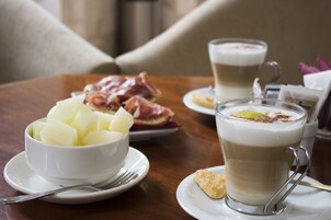 Una mesa con un bol blanco de papas en cubos, una taza de café con una galleta, y un plato con jamón en lonchas.
