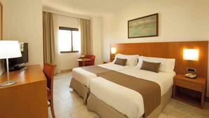 A hotel room with a large bed, a desk with a lamp, a TV, a chair, and a window with curtains.