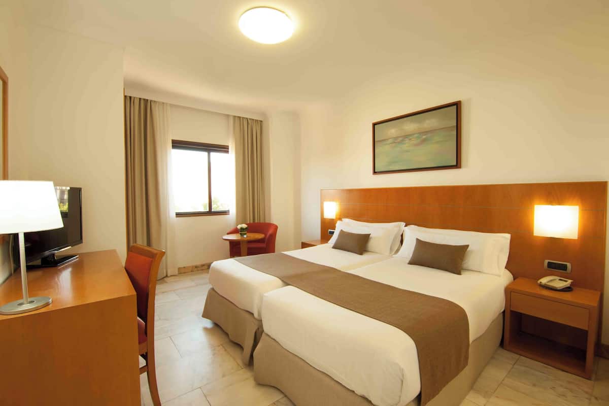 A hotel room with a large bed, a desk with a lamp, a TV, a chair, and a window with curtains.