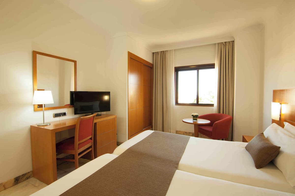 A hotel room with a bed, a desk with a chair, a television, a small table with a chair, and a window with curtains.