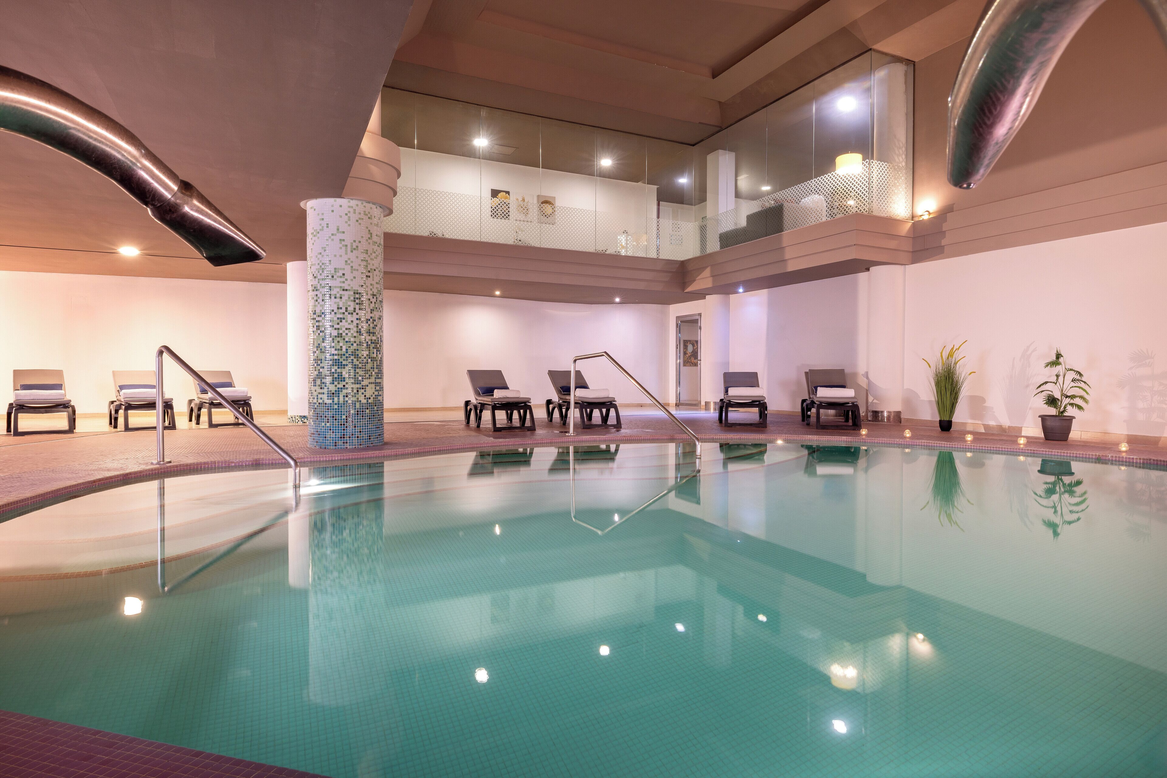 A large indoor swimming pool with clear water, surrounded by lounge chairs and a glass balustrade.