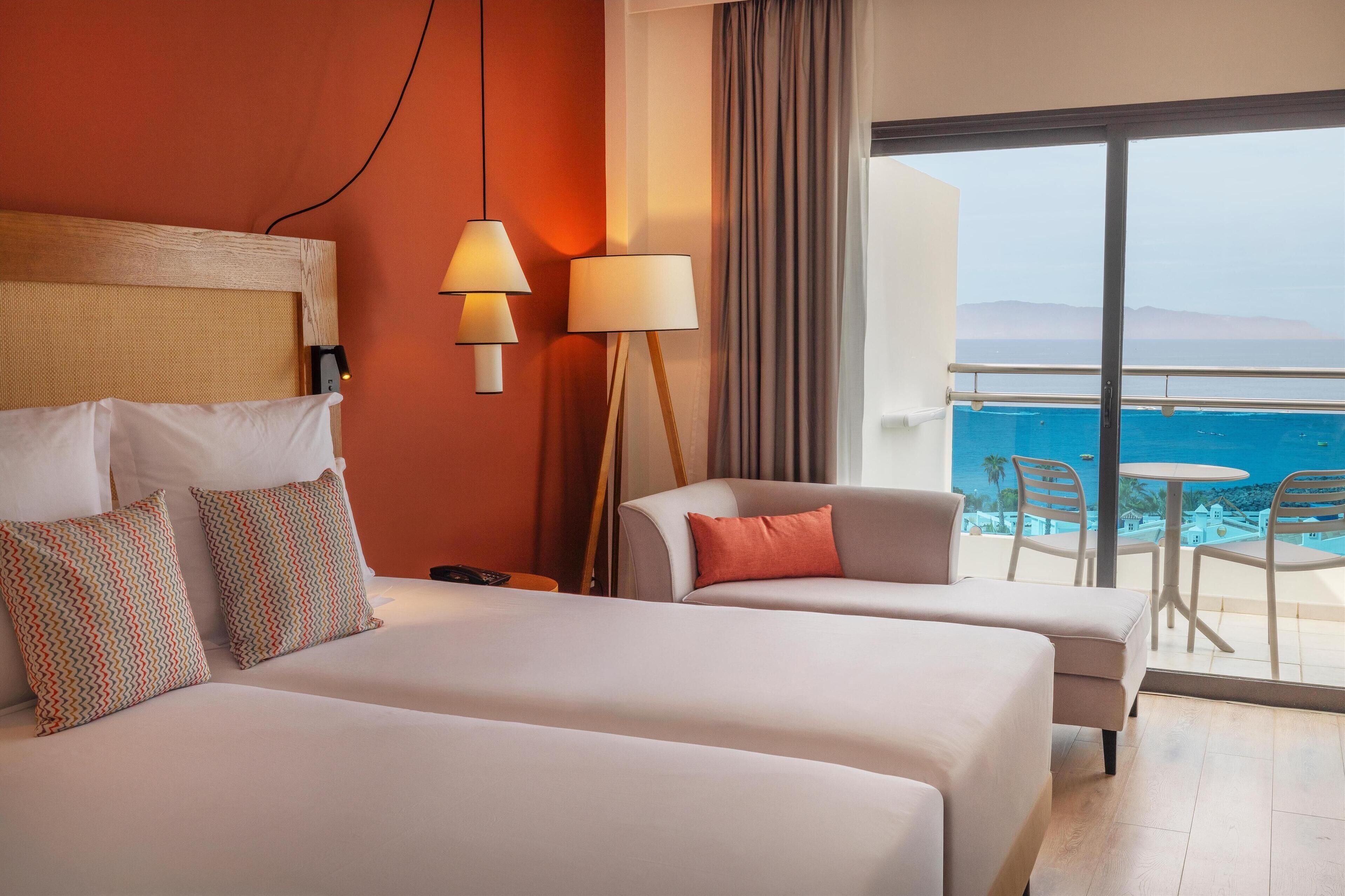 A hotel room with a bed, two bedside lamps, a sofa, and a view of the sea.