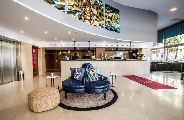 A modern lobby with a curved ceiling, a seating area with a tufted sofa and ottoman, a bar area, and a large window offering a view of the street. - Leonardo Hotel Barcelona Gran Via (Barcelona)