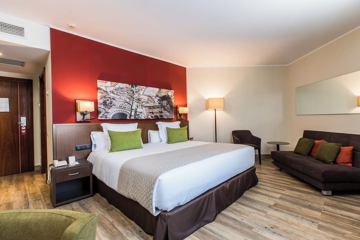 Camera d'albergo con un letto grande, una testiera in legno, una parete di accento rosso, un quadro, un divano e un tavolino.