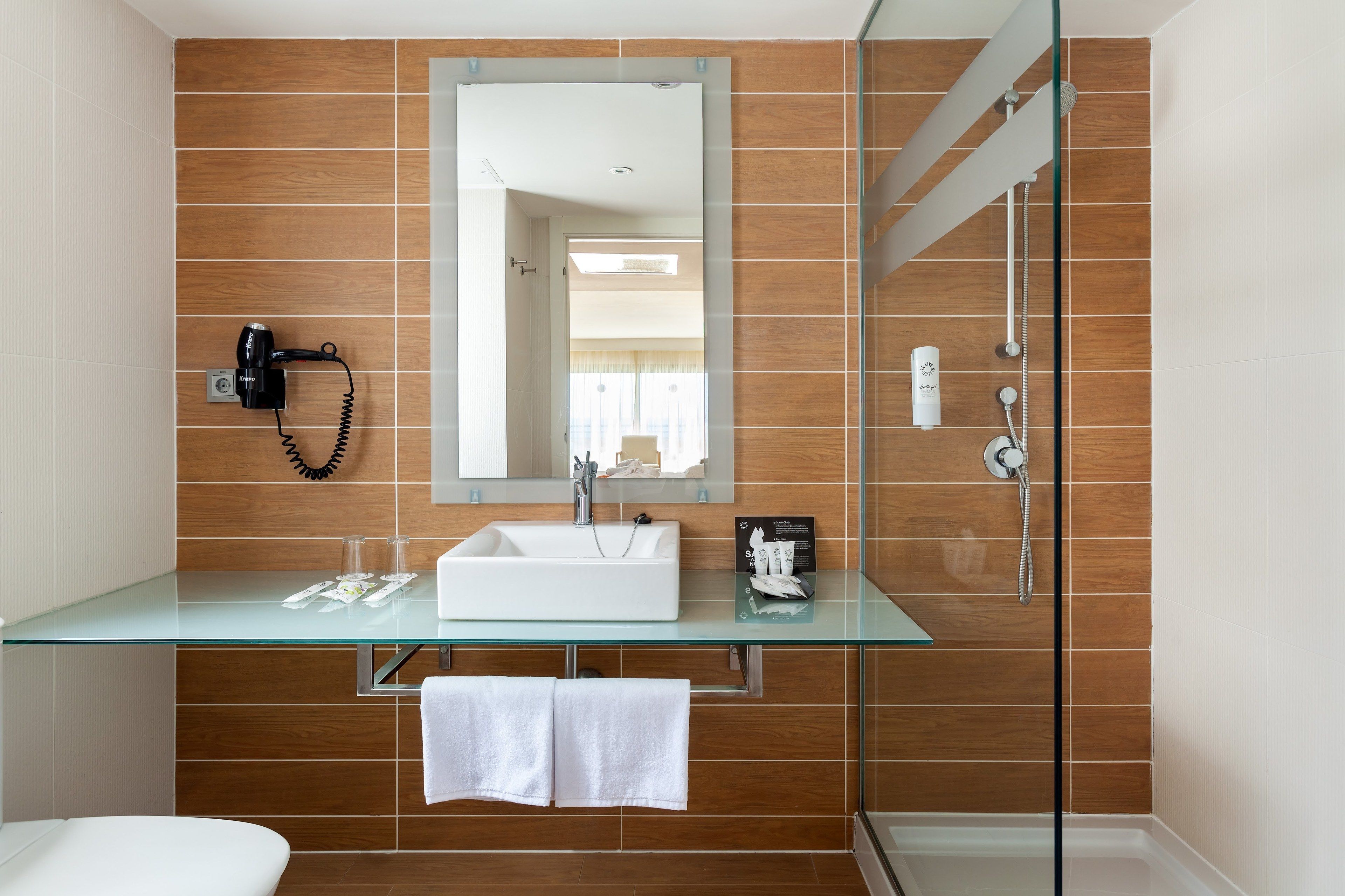 A modern bathroom with a glass vanity, a large mirror, a white sink, a hairdryer, and a shower area.