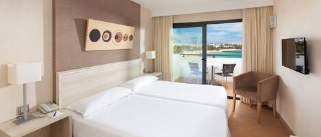 A hotel room with a large bed, a desk with a phone, a chair, a TV, and a view of a pool and beach.