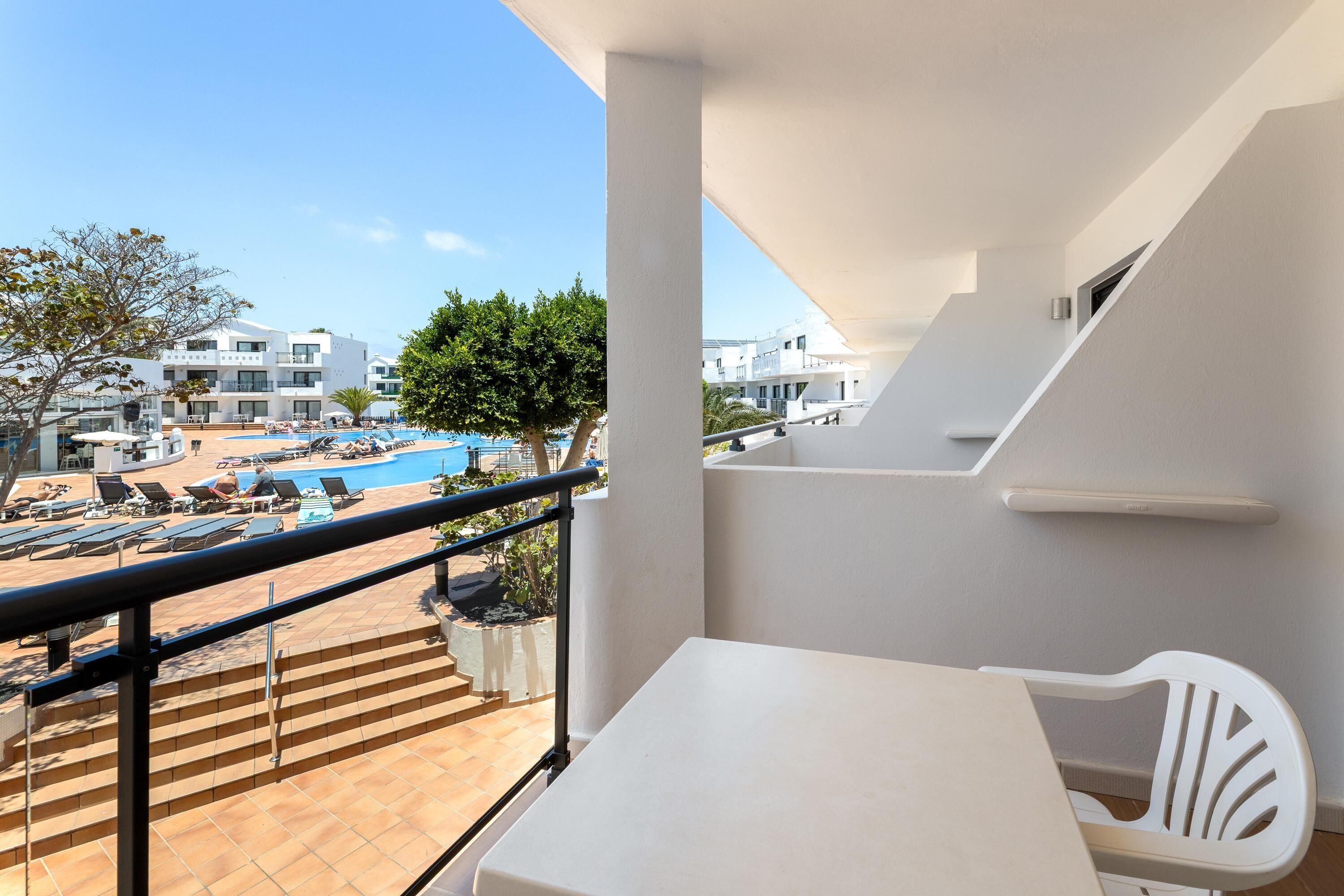 Un balcone con tavolo e sedia bianchi, una piscina sullo sfondo e vista su altri edifici.