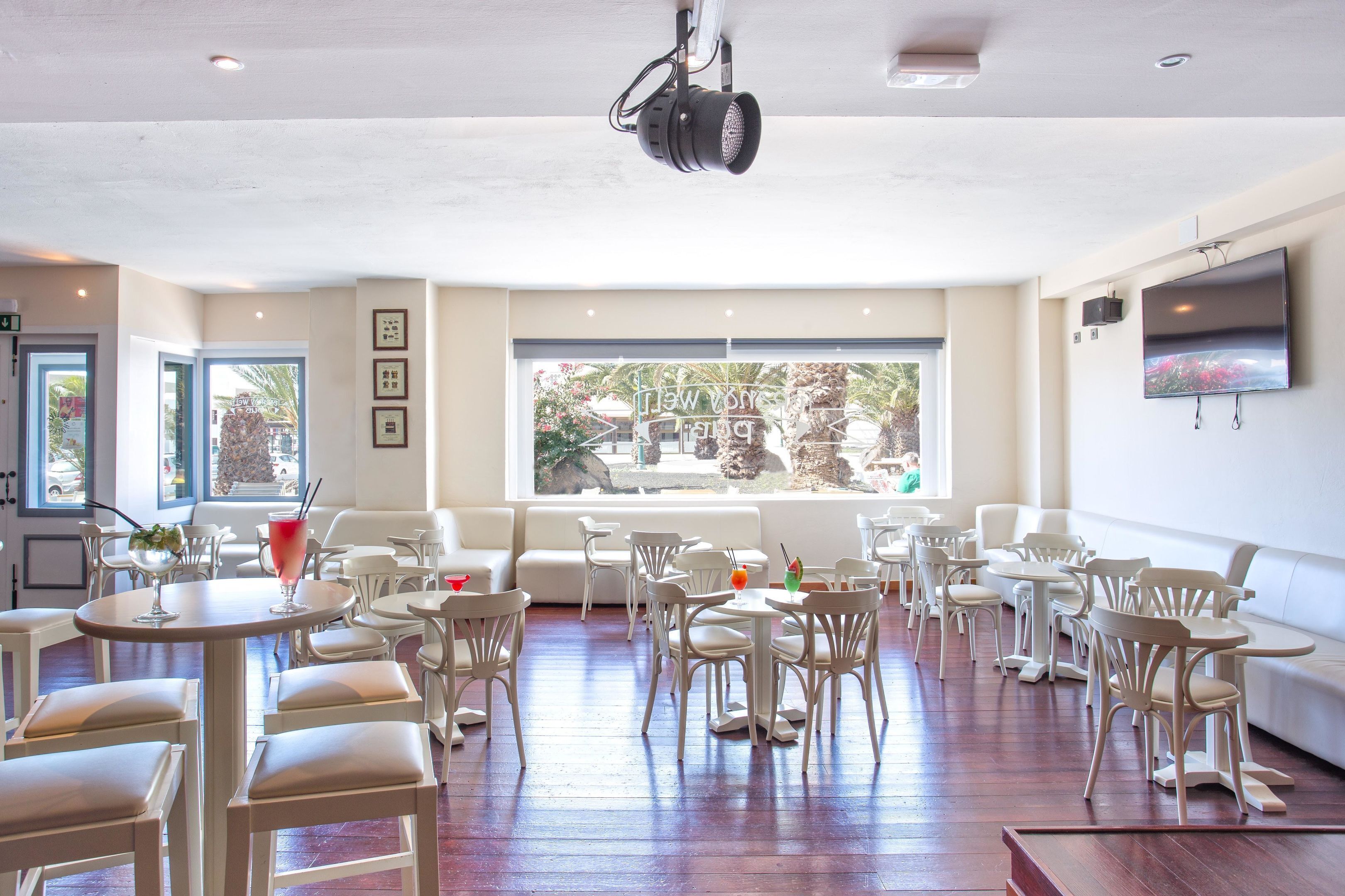 Un vaste espace intérieur aménagé en salle à manger, avec de nombreuses tables et chaises, de grandes fenêtres offrant une vue sur l’extérieur et un téléviseur fixé au mur.