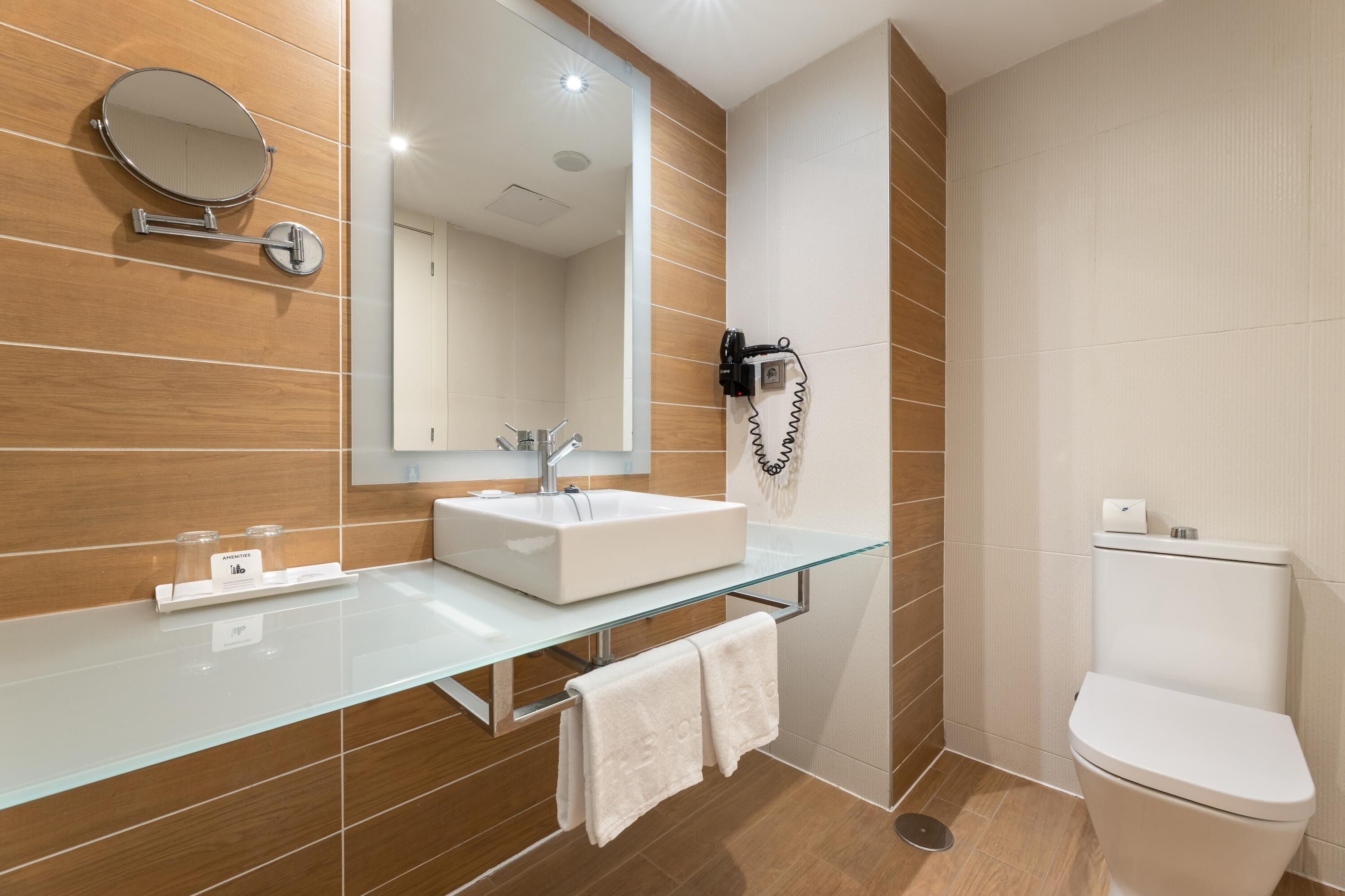 A modern bathroom with a glass vanity, a round mirror, a handheld showerhead, and a toilet.
