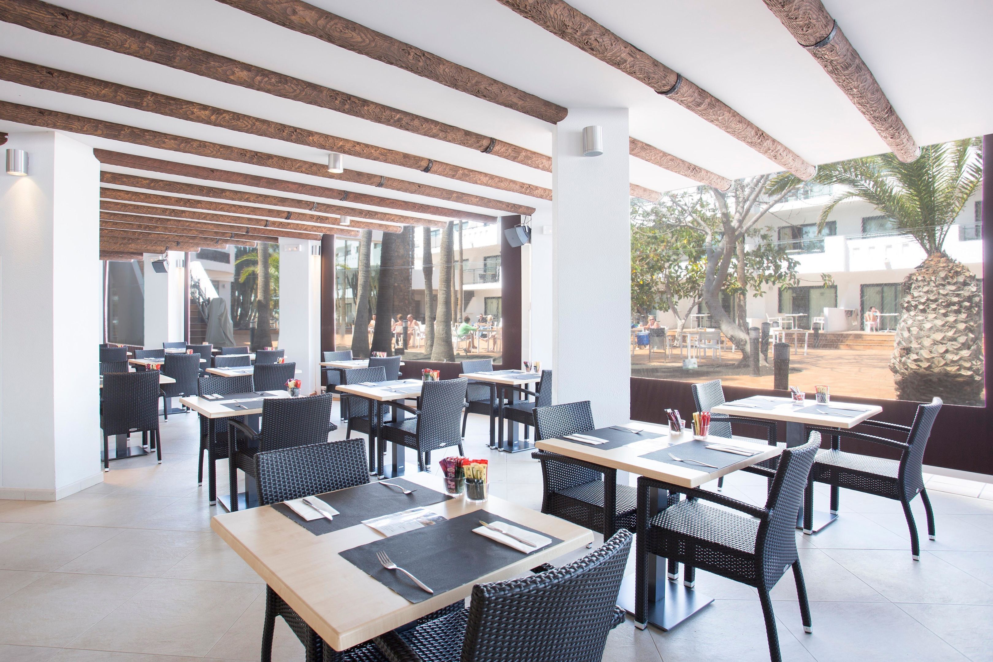 Un amplio espacio exterior para comer con vigas de madera, sillas de mimbre negro y mesas dispuestas para dos.
