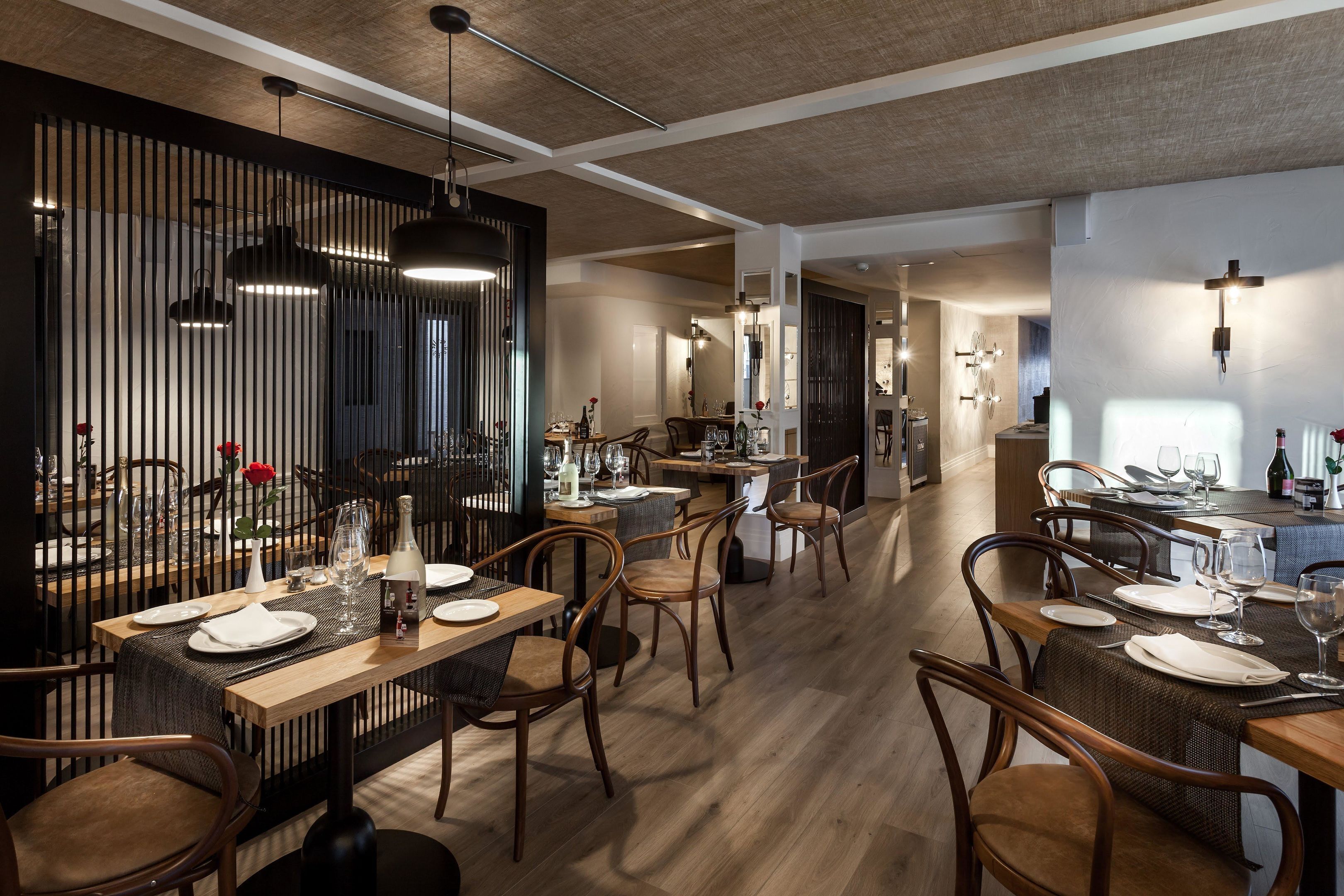 Un comedor moderno con mesas y sillas de madera, una separación oscura y lámparas colgantes.