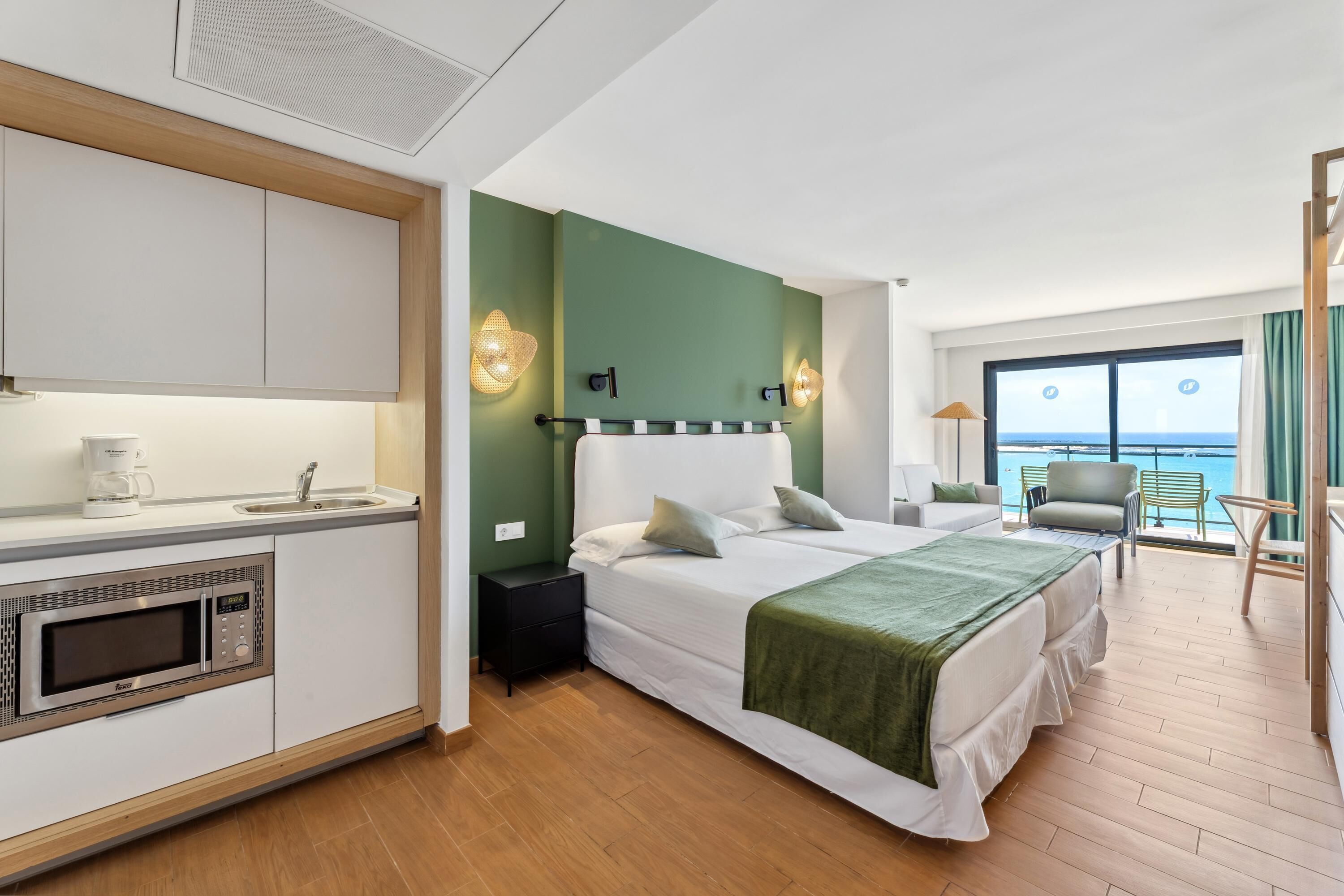 Habitación de hotel moderna con cama, kitchenette, vista al mar y balcón con una silla.