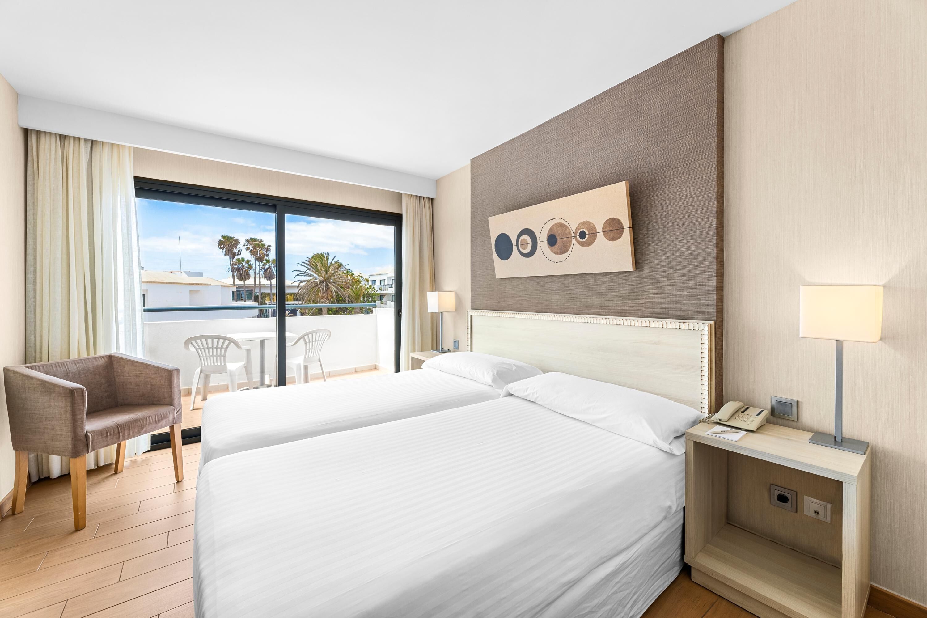 A hotel room with two beds, a desk, a chair, and a view of palm trees and buildings.