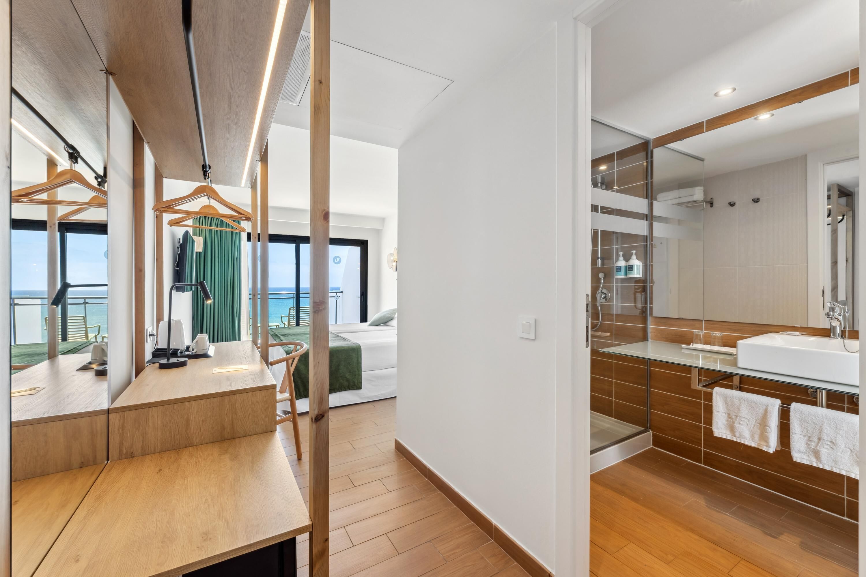Una habitación de hotel moderna con un escritorio de madera, un espejo grande, una cama con cortina verde y un baño con ducha de vidrio y lavamanos blanco.