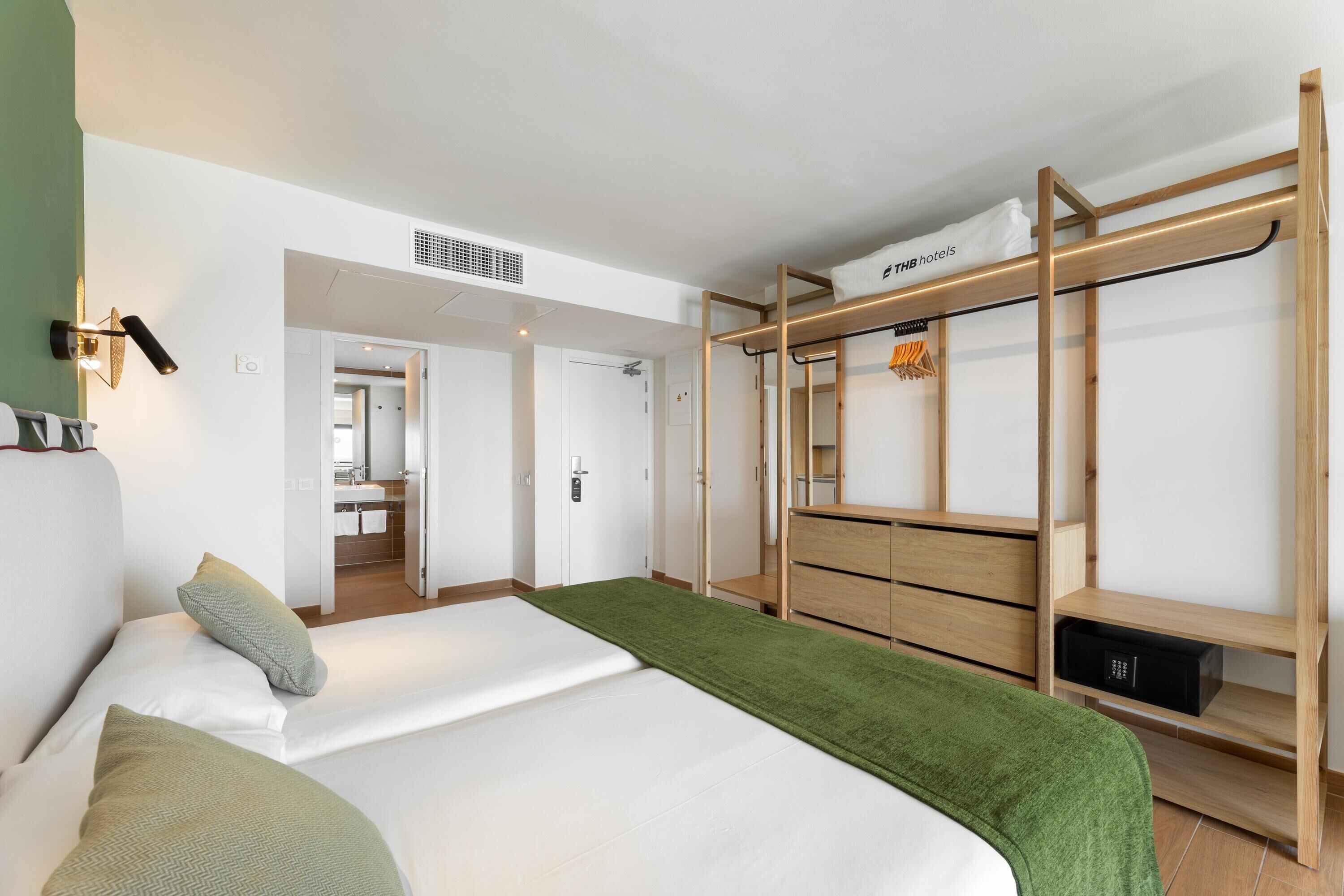 Une chambre d’hôtel moderne, équipée d’un lit, d’une armoire en bois, d’un miroir et d’une salle de bain visible à travers une porte ouverte.