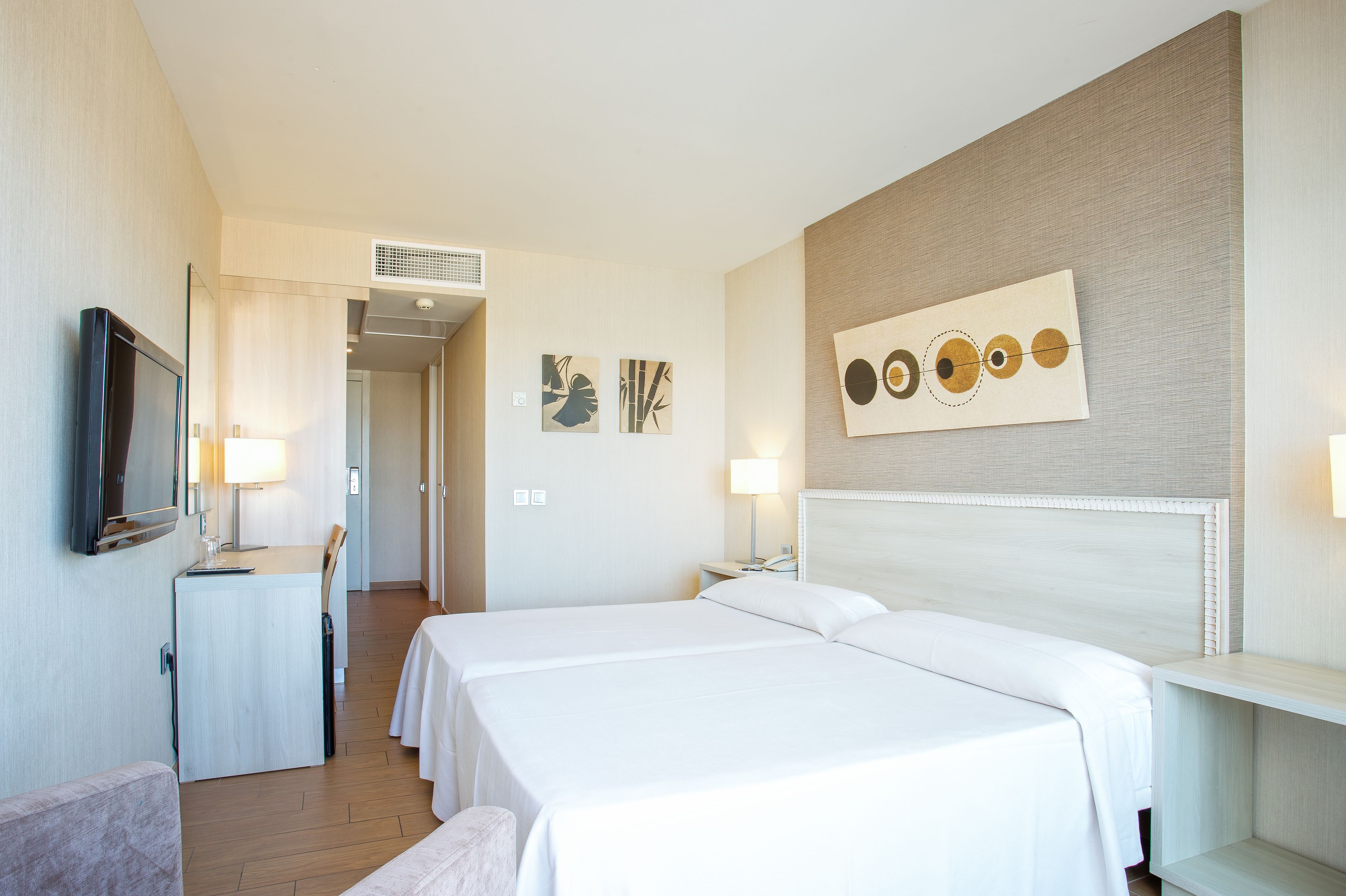 A hotel room with two beds, a desk, a chair, a TV, and artwork on the walls.