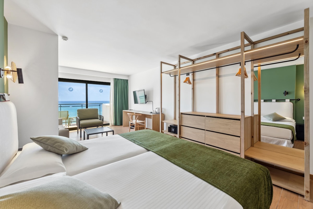 Ett hotellrum med två sängar, ett skrivbord, en stol, en garderob och en balkong med utsikt över havet.