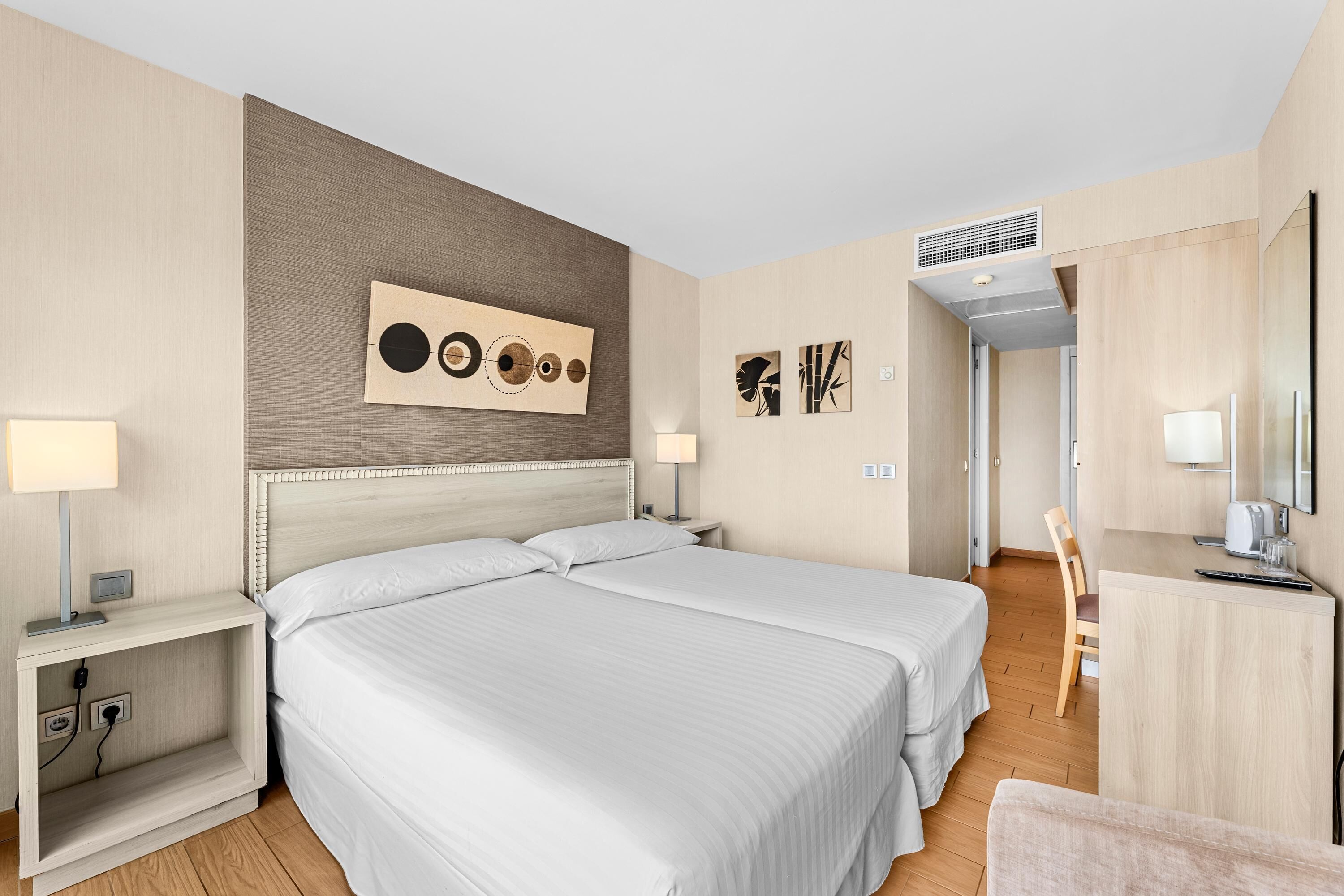 Habitación de hotel con dos camas, un escritorio y una silla. Se observan obras de arte en la pared y un respiradero en el techo.
