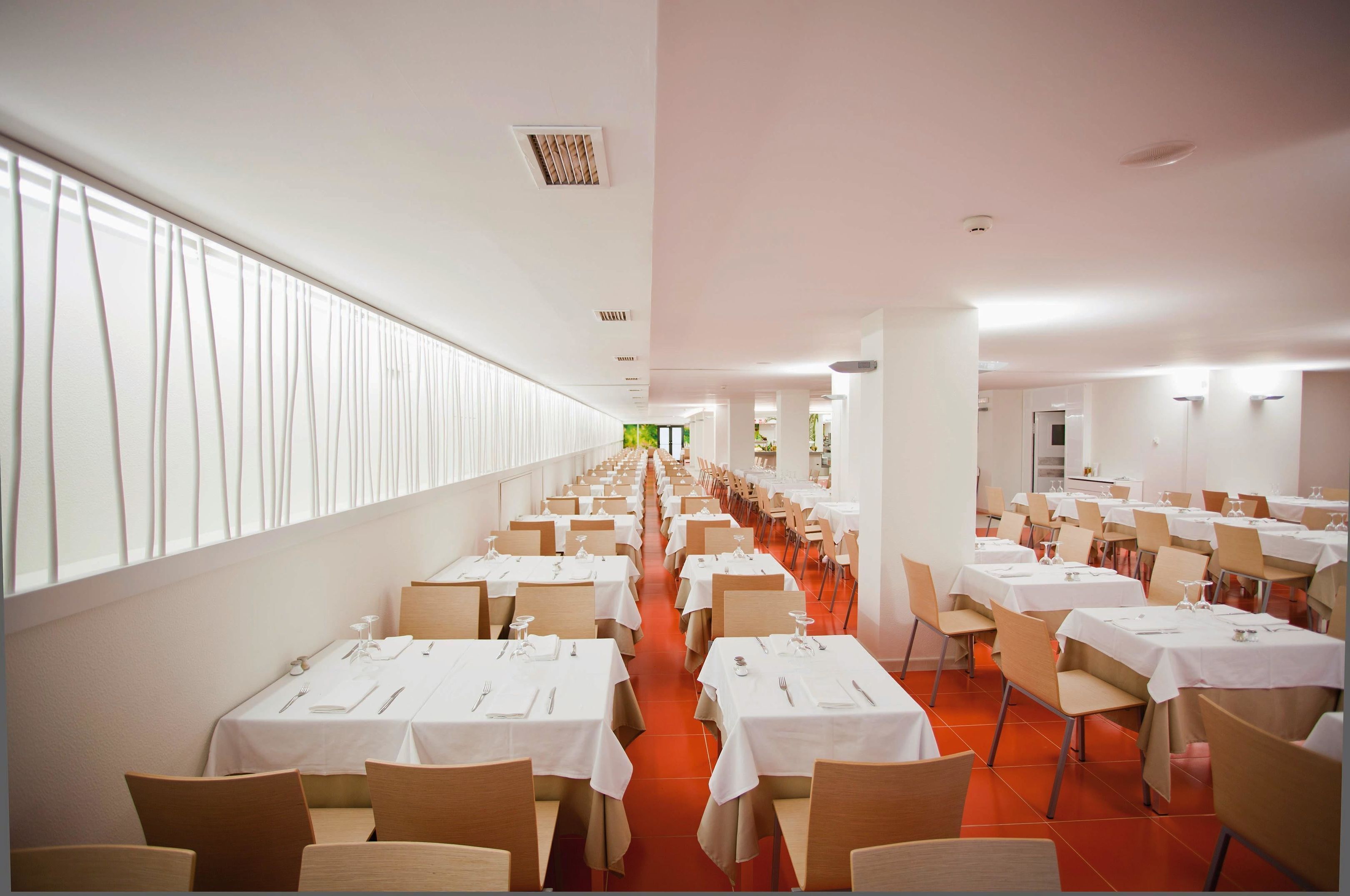 Un comedor amplio con filas de mesas y sillas, ventanas grandes a lo largo de una pared, y un ambiente luminoso.