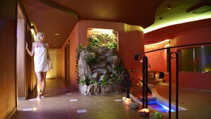 Un'area benessere con pavimento in legno, parete rocciosa con piante e vasca idromassaggio illuminata.