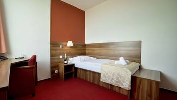 Ett hotellrum med en sänggavel i trä, en säng med vita sängkläder, en röd matta, ett skrivbord med en stol och ett nattduksbord med en lampa.