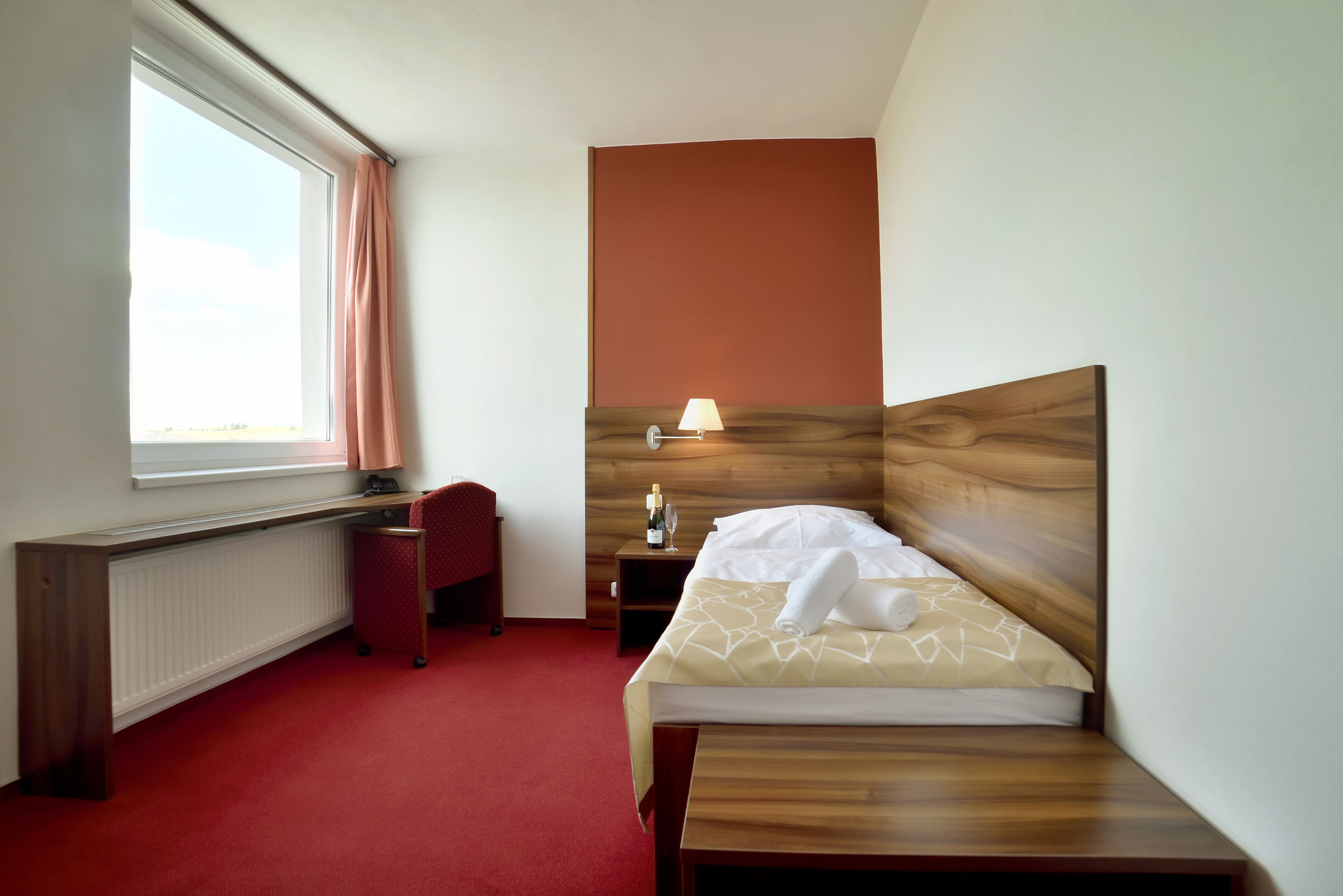 A hotel room with a bed, a desk, a chair, a nightstand, and a lamp.