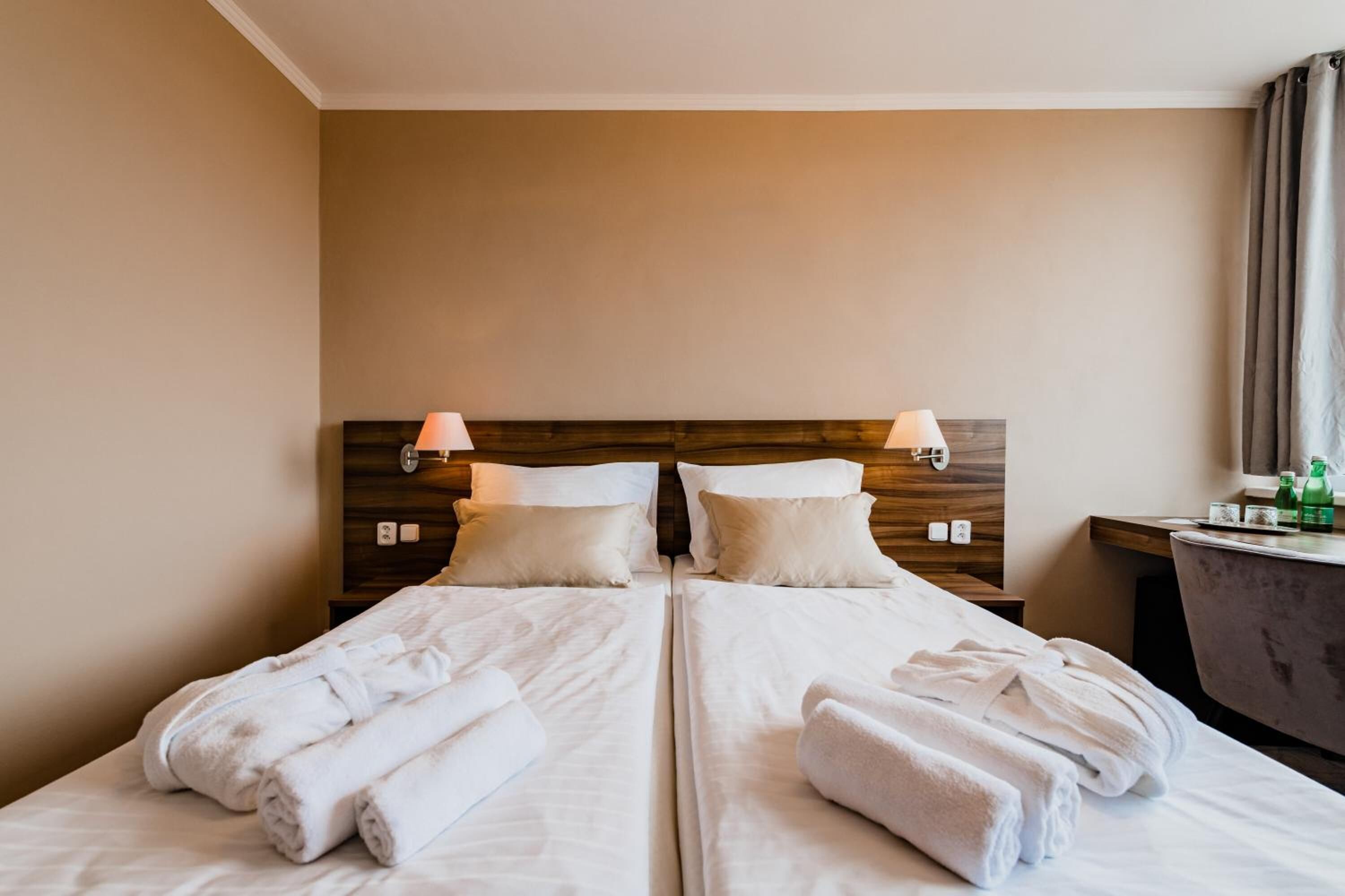 A double bed with white linens and towels, a wooden headboard, bedside lamps, and a small table with toiletries.