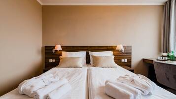 A double bed with white linens and towels, a wooden headboard, bedside lamps, and a small table with toiletries.