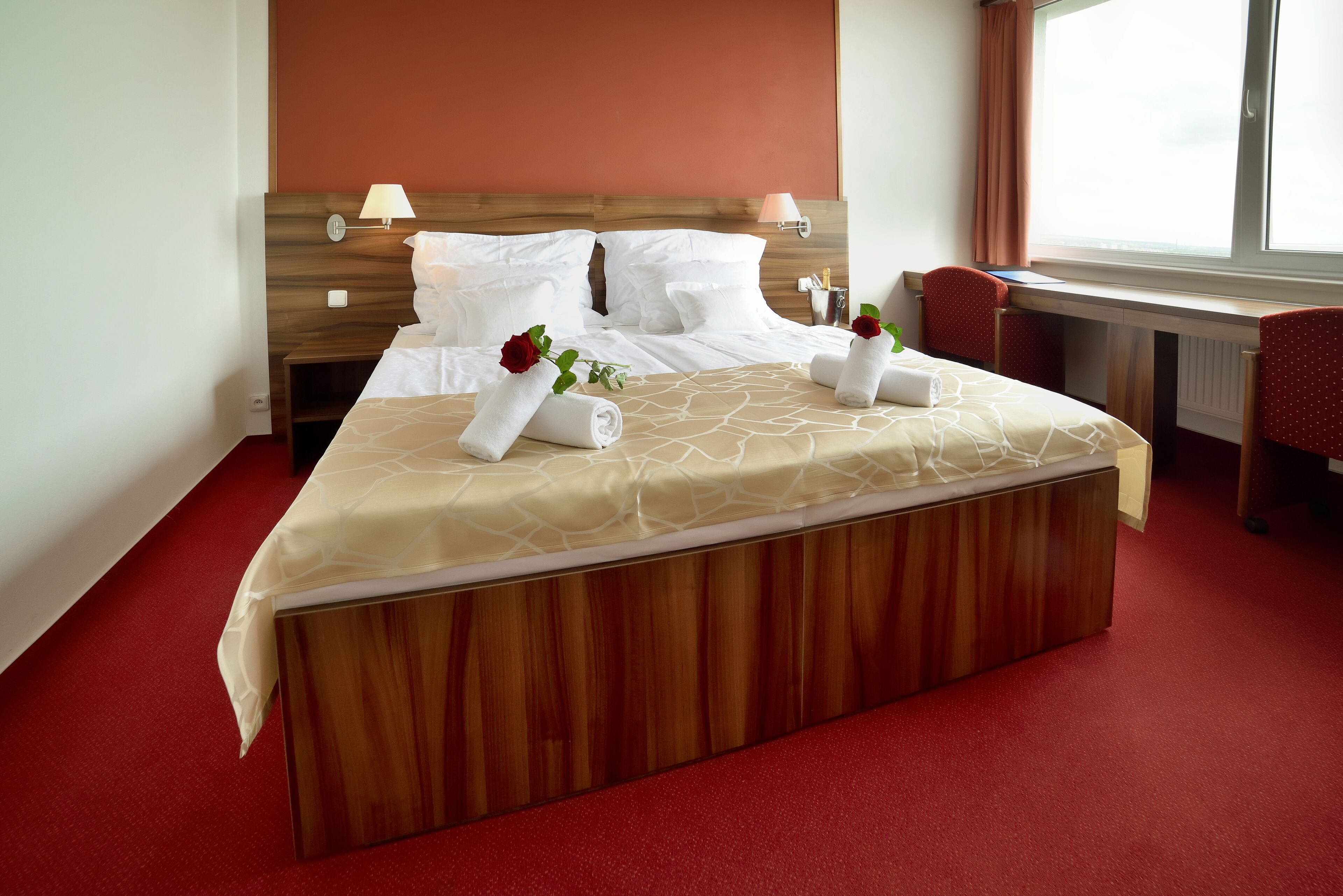 A neatly made bed with white linens, two red roses, and a wooden headboard.