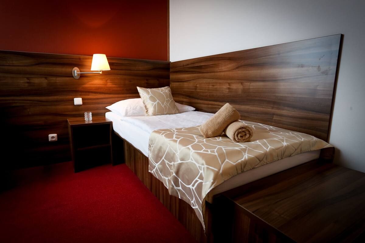 Een hotelkamer met een houten hoofdbord, een rood tapijt, een nachtkastje met een lamp, en een bed met een geborduurde spreideken en kussens.