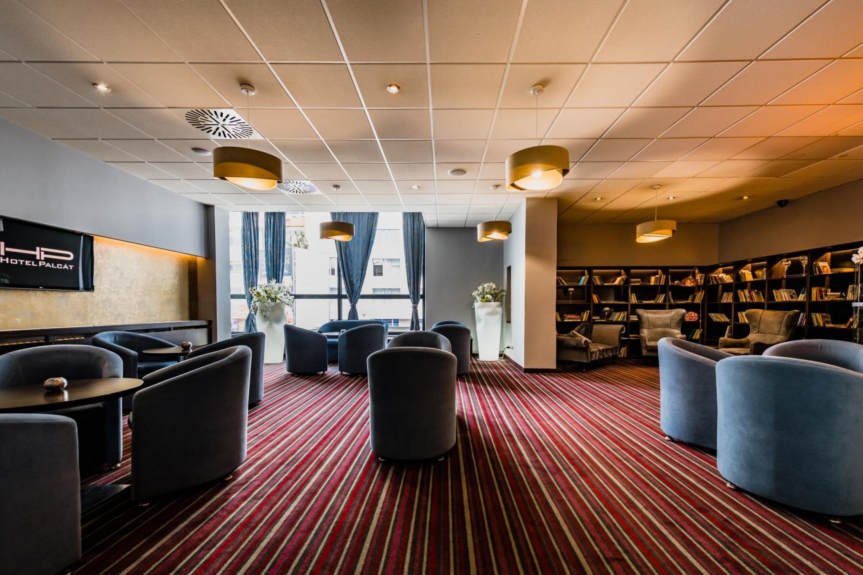 A hotel lobby with a red striped carpet, seating areas with armchairs and tables, a bookshelf, and a large window with curtains.