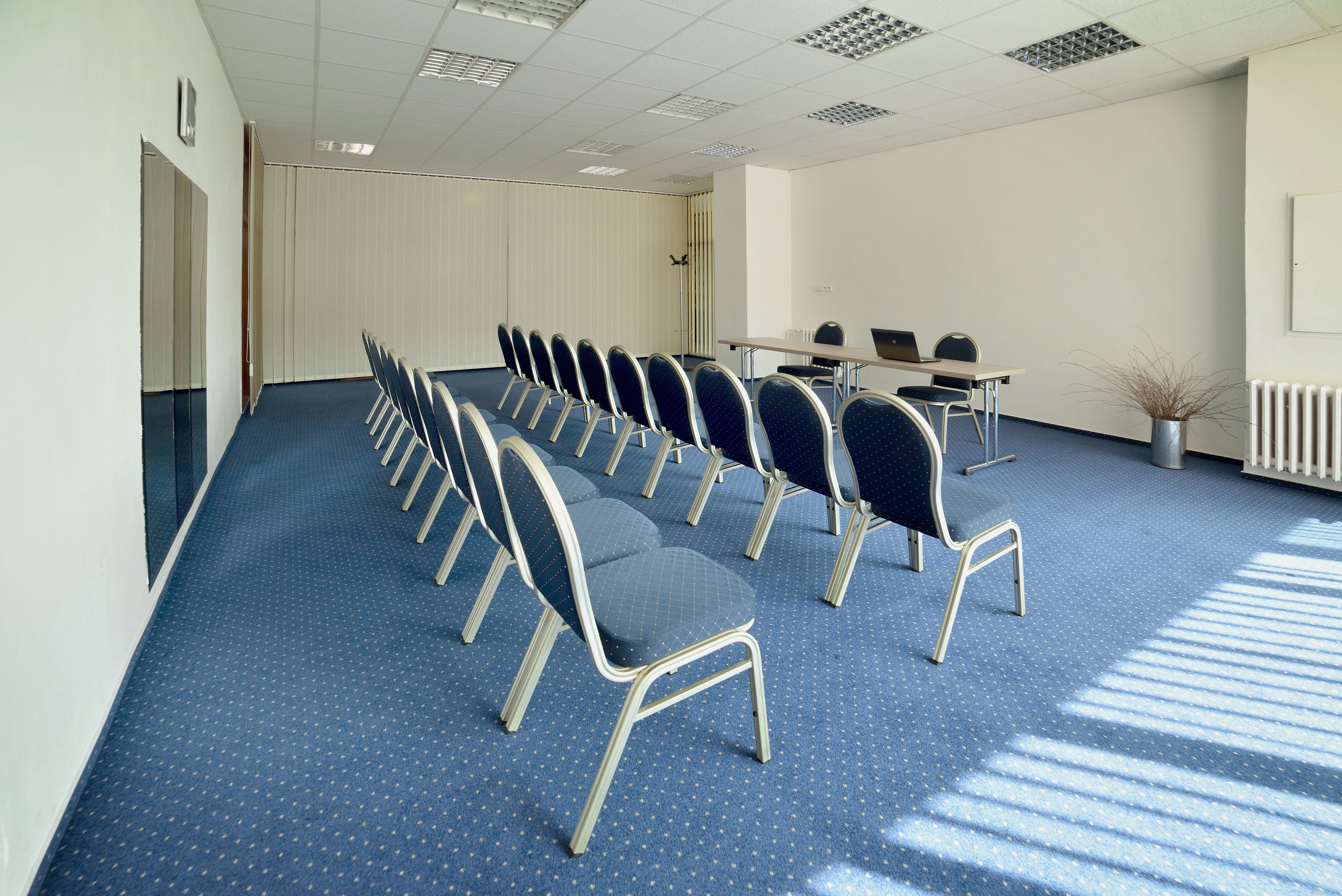 Un salón de conferencias con filas de sillas, una mesa con portátiles y un radiador.