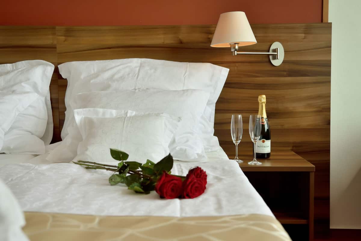 Een net opgemaakt bed met witte lakens, een boeket rode rozen en een fles champagne op een nachtkastje van hout.