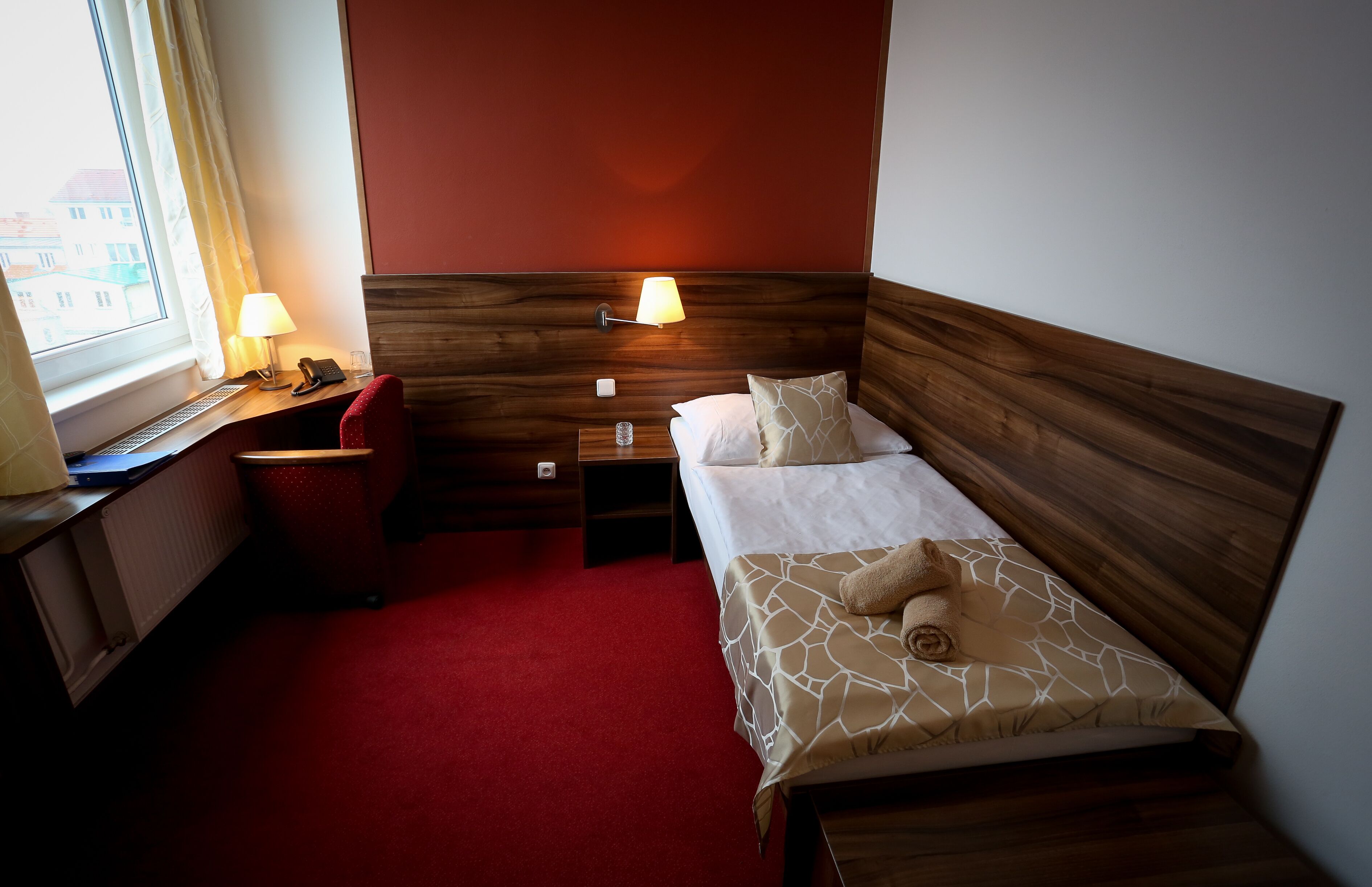 A hotel room with a wooden headboard, a red carpet, a desk with a lamp, a telephone, and a window with curtains.
