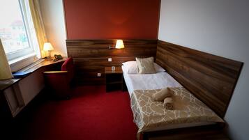 A hotel room with a wooden headboard, a red carpet, a desk with a lamp, a telephone, and a window with curtains.