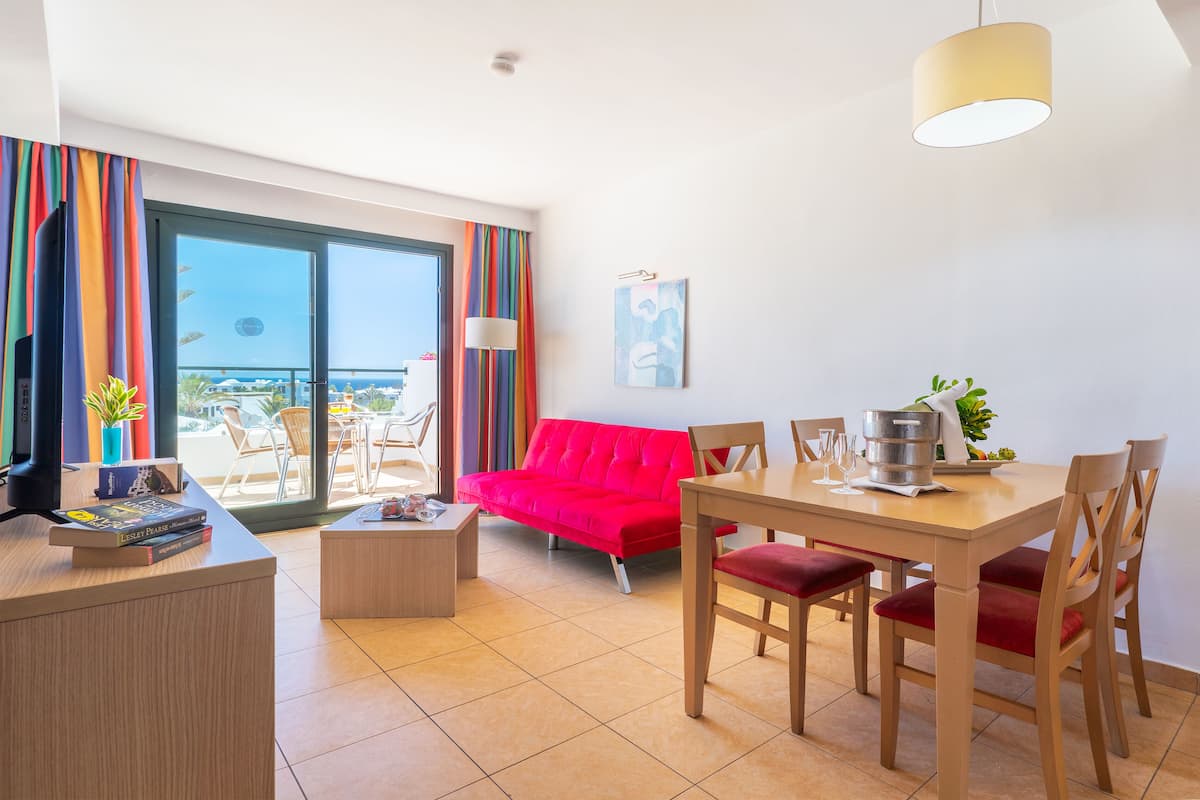 A modern hotel room with a balcony, a red sofa, a wooden dining table with chairs, and a desk with books and a lamp.
