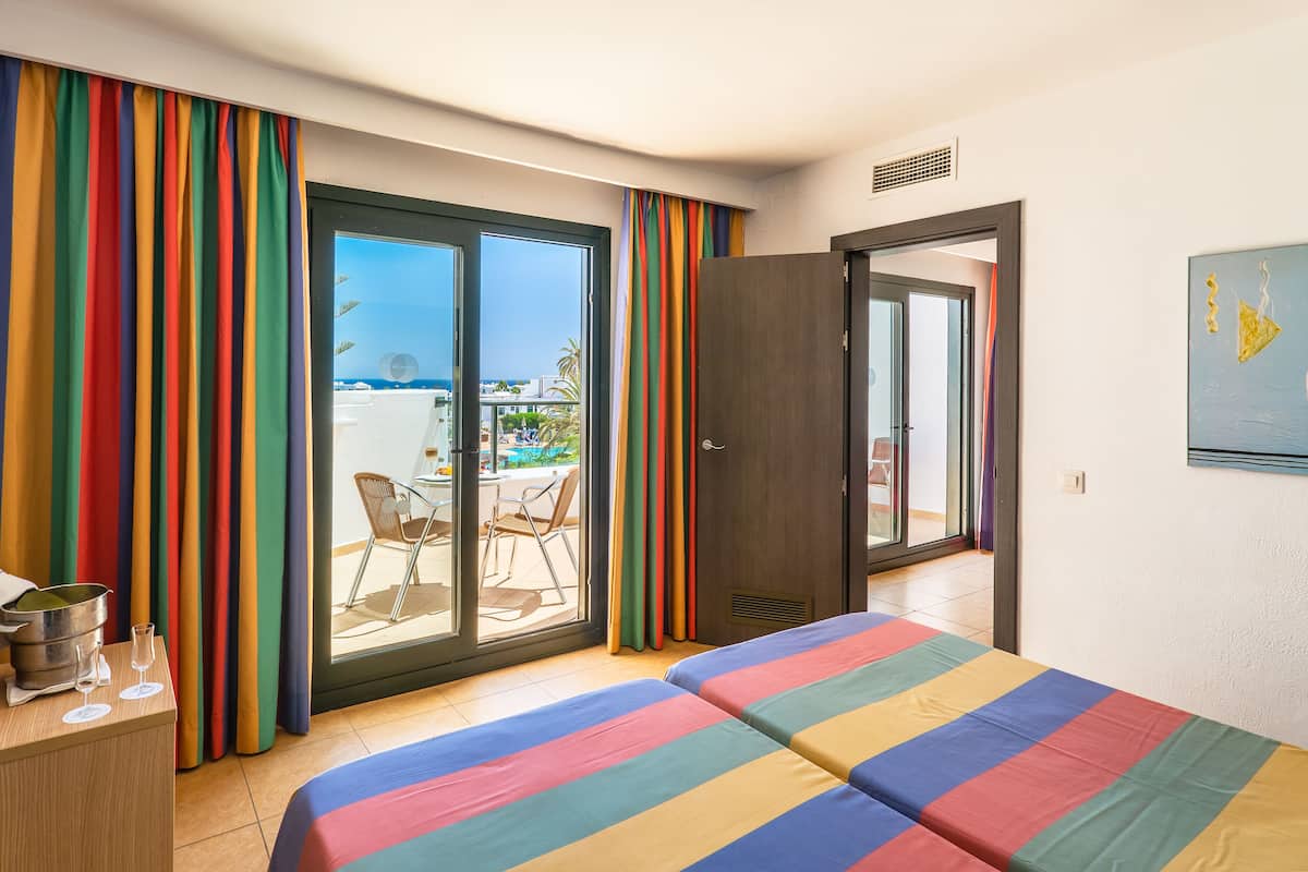 A hotel room with a large bed, a desk with a kettle and glasses, and a view of a balcony with outdoor furniture.